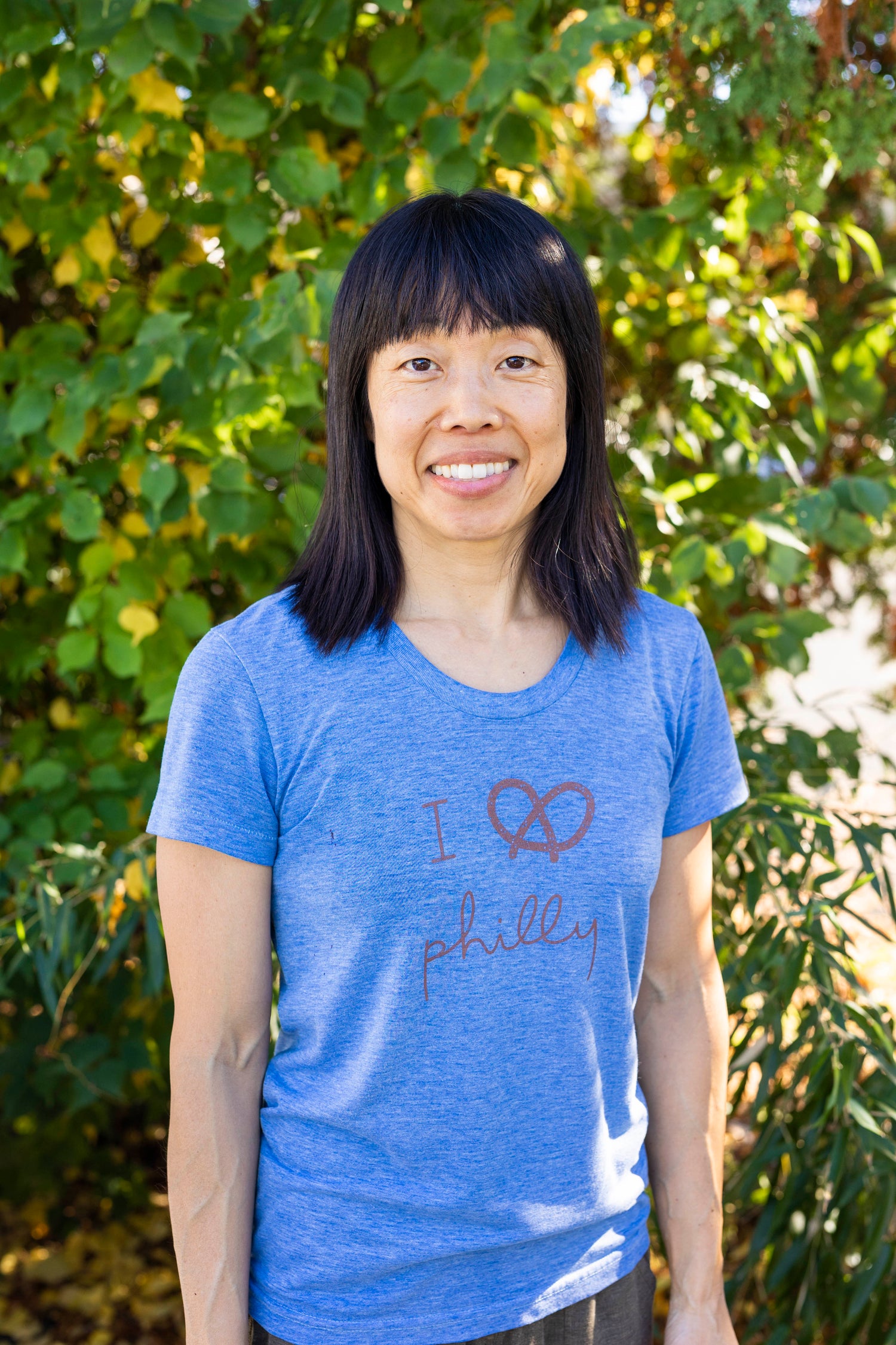 Photo of smiling woman with medium length dark hair in blue t-shirt with trees in background.
